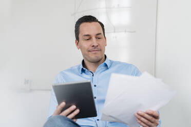 Male entrepreneur holding digital tablet and documents while sitting at office - DIGF14981