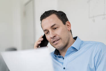 Männlicher Unternehmer, der mit einem Smartphone spricht und ein Dokument im Büro hält - DIGF14984