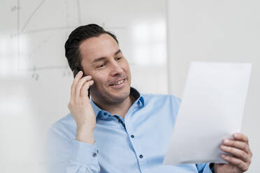 Männlicher Berufstätiger, der am Arbeitsplatz mit einem Smartphone telefoniert und ein Dokument in der Hand hält - DIGF14985