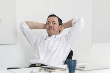Thoughtful male professional with hands behind head sitting at workplace - DIGF14996