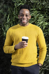 Happy man with hand in pocket holding disposable coffee cup in front of lush foliage - FBAF01768