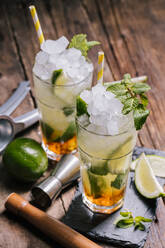 Hoher Winkel von Gläsern mit kaltem Alkohol Mojito-Cocktail mit Rum und frischer Limette, serviert mit Eis und Minzblättern auf Holztisch mit Auspresser und Jigger - ADSF22450