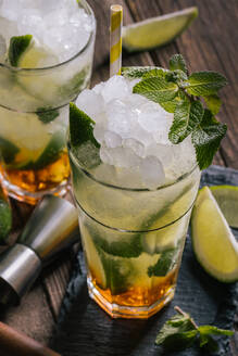 High angle of glasses with cold alcohol Mojito cocktail with rum and fresh lime served with ice and mint leaves on wooden table with squeezer and jigger - ADSF22451