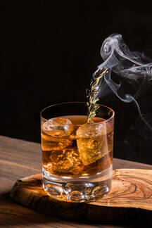 https://us.images.westend61.de/0001543132i/glass-of-alcohol-old-fashioned-whiskey-cocktail-with-ice-cubes-and-fuming-rosemary-branch-on-wooden-table-ADSF22455.jpg