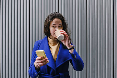Female entrepreneur drinking coffee while using smart phone in front of wall - ASGF00096