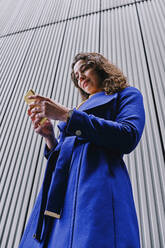 Geschäftsfrau im Trenchcoat, die ein Mobiltelefon benutzt, während sie vor einer grauen Wand steht - ASGF00101