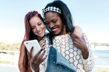 Happy young multiracial lesbian couple hugging and browsing mobile phone together while spending summer day on seashore - ADSF22464