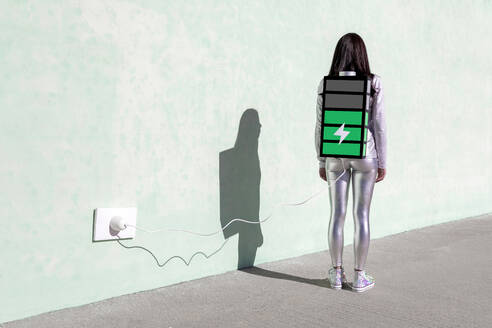 Back view of unrecognizable female with long dark hair in stylish outfit standing on street near wall with battery shaped backpack connected to charger - ADSF22466