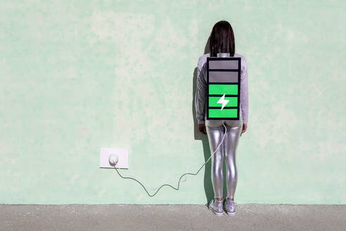 Rückenansicht einer nicht erkennbaren Frau mit langen dunklen Haaren in stilvollem Outfit, die auf der Straße in der Nähe einer Mauer steht und einen Rucksack in Form einer Batterie trägt, die an ein Ladegerät angeschlossen ist - ADSF22471