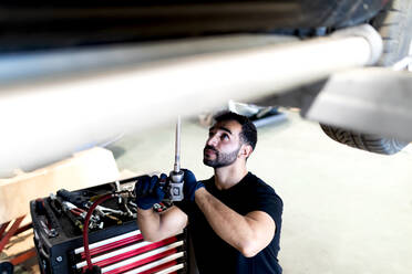 Beschäftigter männlicher Techniker, der ein spezielles Instrument benutzt und ein Auto repariert, während er in einer modernen Werkstatt arbeitet - ADSF22472