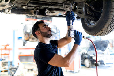 Beschäftigter männlicher Techniker, der ein spezielles Instrument benutzt und ein Auto repariert, während er in einer modernen Werkstatt arbeitet - ADSF22473