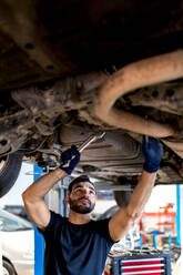Low angle of bearded male mechanic with wrench standing under car and inspecting detail with metal wrench - ADSF22474