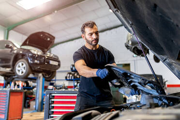 Low angle of busy male master taking off plastic protective cover from car engine while fixing vehicle in workshop - ADSF22476