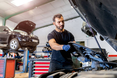 Niedriger Winkel des beschäftigten männlichen Meisters, der die Plastikschutzabdeckung vom Automotor abnimmt, während er das Fahrzeug in der Werkstatt repariert - ADSF22476