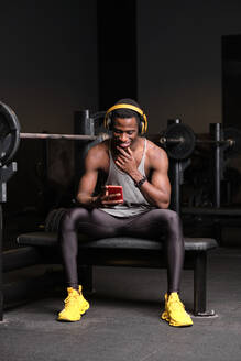 Lächelnder junger afrikanischer Mann mit Kopfhörern und Smartphone im Fitnessstudio - AODF00445