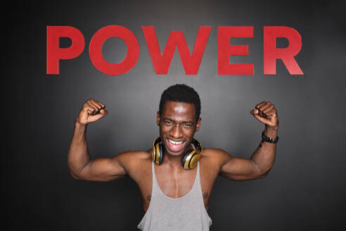 Männlicher Sportler, der seine Muskeln unter dem Powertext vor einer Wand im Fitnessstudio anspannt - AODF00456