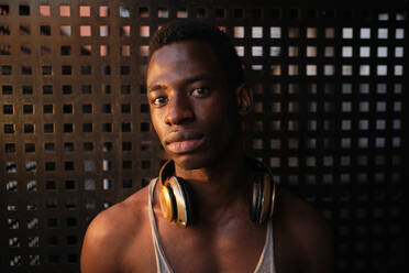 Junger Mann mit Kopfhörern um den Hals vor einer Metallwand im Fitnessstudio - AODF00458