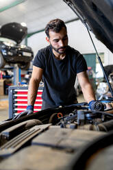 Seriöser männlicher Mechaniker, der den Deckel eines Automotors öffnet und das Öl während einer Untersuchung im Service überprüft - ADSF22477