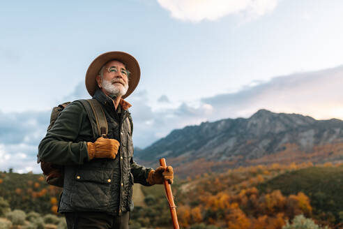 Seitenansicht eines älteren männlichen Wanderers mit Holzstock, der auf einem Hügel steht und die malerische Aussicht auf das Hochland von Cáceres bewundert - ADSF22480