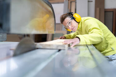 Seitenansicht einer fokussierten erwachsenen Handwerkerin mit Schutzbrille und Kopfhörern, die mit einer scharfen Kreissäge in einer Werkstatt ein Holzbrett sägt - ADSF22485