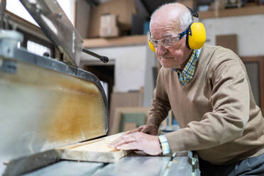 Focused adult elderly man artisan in goggles and headphones sawing wooden board with sharp circular saw in workshop - ADSF22487