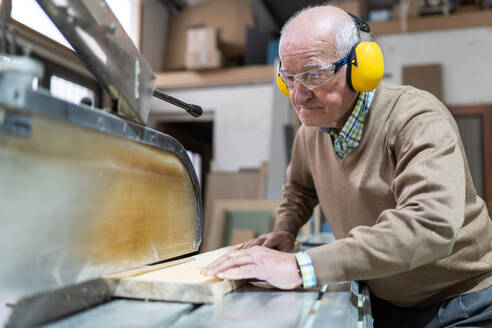 Fokussierter erwachsener älterer Mann mit Schutzbrille und Kopfhörern sägt ein Holzbrett mit einer scharfen Kreissäge in einer Werkstatt - ADSF22487