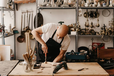 Holzarbeiter in Schürze beim Schnitzen von Holz mit Meißel und Hammer, während er in der Werkstatt kunstvolle Details anfertigt - ADSF22492