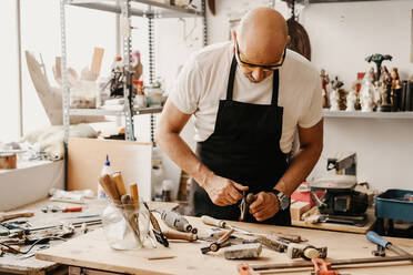 Focused mature woodworker choosing instruments from wooden workbench while working in creative workshop - ADSF22493