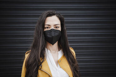Mid adult woman with protective face mask standing in front of black shutter - EBBF03077