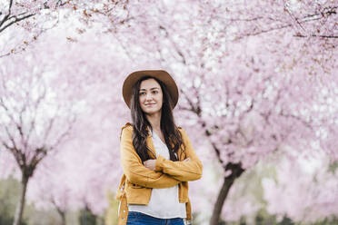 Beautiful woman with arms crossed looking away while standing in front of almond trees - EBBF03081
