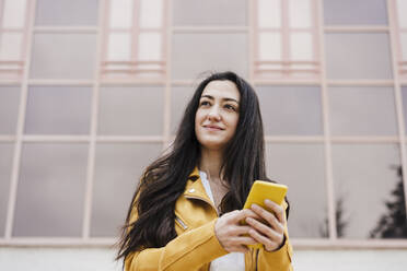 Smiling woman looking away while using mobile phone in front of building - EBBF03088