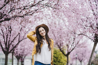 Smiling woman wearing sun hat while standing in front of trees during springtime - EBBF03094