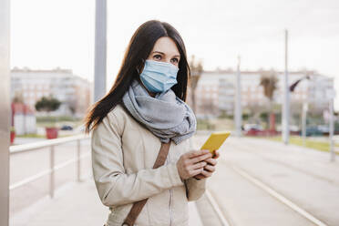 Mid adult woman with protective face mask looking away while holding mobile phone at railroad station - EBBF03107