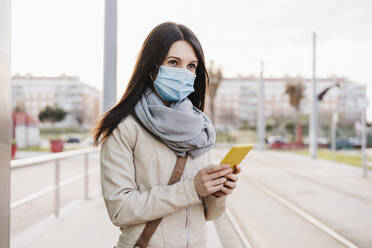 Mittlere erwachsene Frau mit Schutzmaske, die wegschaut, während sie ein Mobiltelefon am Bahnhof hält - EBBF03107