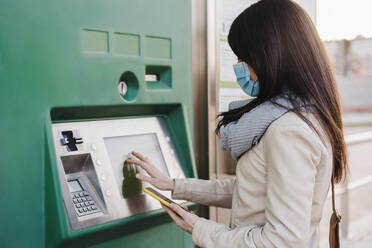 Woman wearing protective face mask operating ticket machine while holding mobile phone during pandemic - EBBF03110