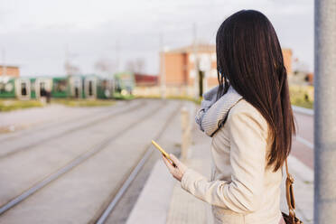 Frau mit langen Haaren, die ihr Smartphone hält, während sie am Bahnsteig auf einen Zug wartet - EBBF03111