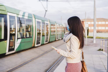 Mid adult woman with mobile phone wearing protective face mask while looking at train on platform - EBBF03112