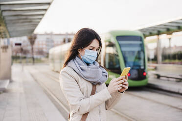 Woman using mobile phone while standing on railway platform during COVID-19 - EBBF03114