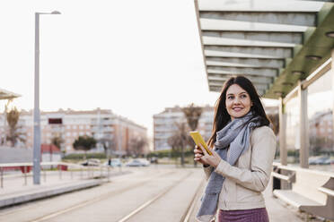 Smiling beautiful woman holding smart phone while looking away at railroad station - EBBF03118