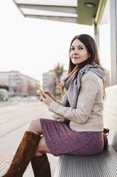 Beautiful woman with smart phone sitting on bench at railroad station - EBBF03120