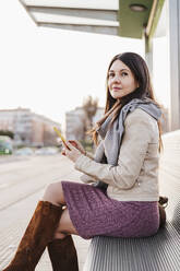 Schöne Frau mit Smartphone sitzt auf Bank am Bahnhof - EBBF03120