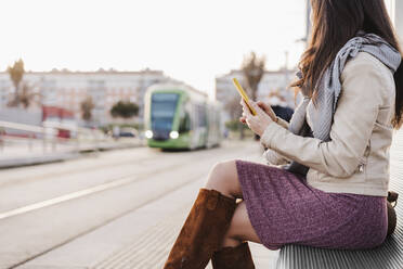 Woman in warm clothing looking at train while sitting with mobile phone on bench - EBBF03121