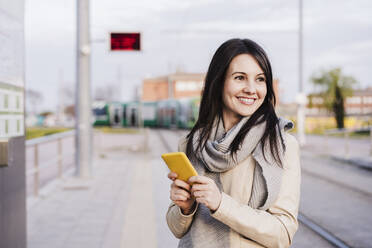 Lächelnde schöne Frau, die mit ihrem Smartphone am Bahnhof steht und nachdenkt - EBBF03122