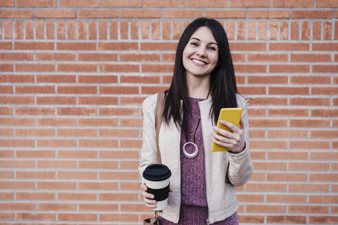 Lächelnde schöne Frau hält Smartphone und Einweg-Kaffeebecher vor einer Backsteinmauer - EBBF03129