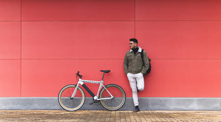 Young businessman by bicycle leaning on red wall - JAQF00446