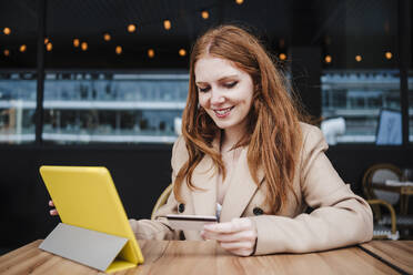 Lächelnde Frau schaut auf ihre Kreditkarte, während sie in einem Café vor einem digitalen Tablet sitzt - EBBF03135