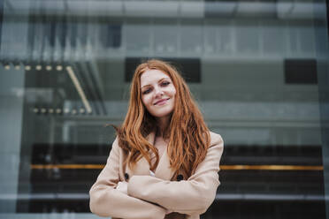 Smiling young woman with arms crossed standing in front of glass - EBBF03163