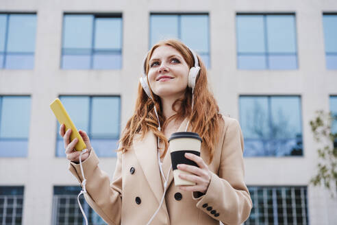 Zufriedene Frau mit Kopfhörern, die ihr Handy und eine Einweg-Kaffeetasse in der Hand hält und wegschaut - EBBF03165