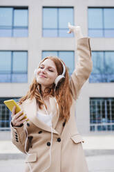 Smiling woman with smart phone listening music through headphones while dancing in front of building - EBBF03166