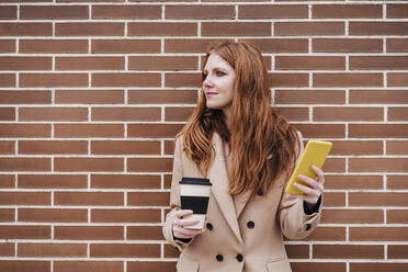 Junge Frau mit rotem Kopf hält Smartphone und Einweg-Kaffeebecher vor einer Wand - EBBF03173